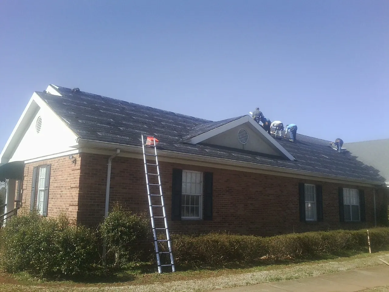 Crew tearing shingles off a brick ranch roof with a ladder set — Emergency Roof Repair work by our crew in Rialto
