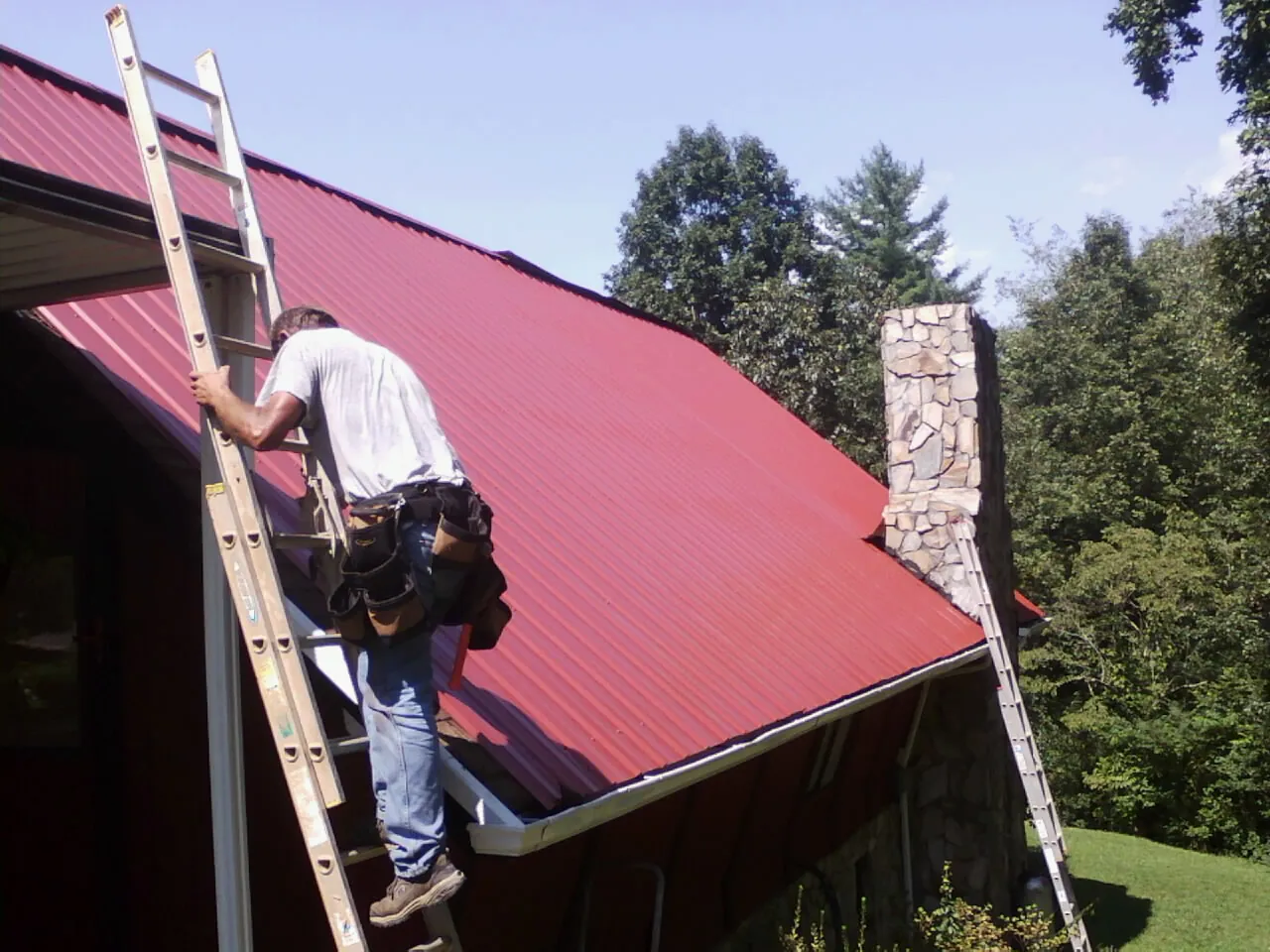 Roofer on a ladder beside a red metal roof and stone chimney — Soffit & Fascia Repair by Roofing Brothers in Rialto